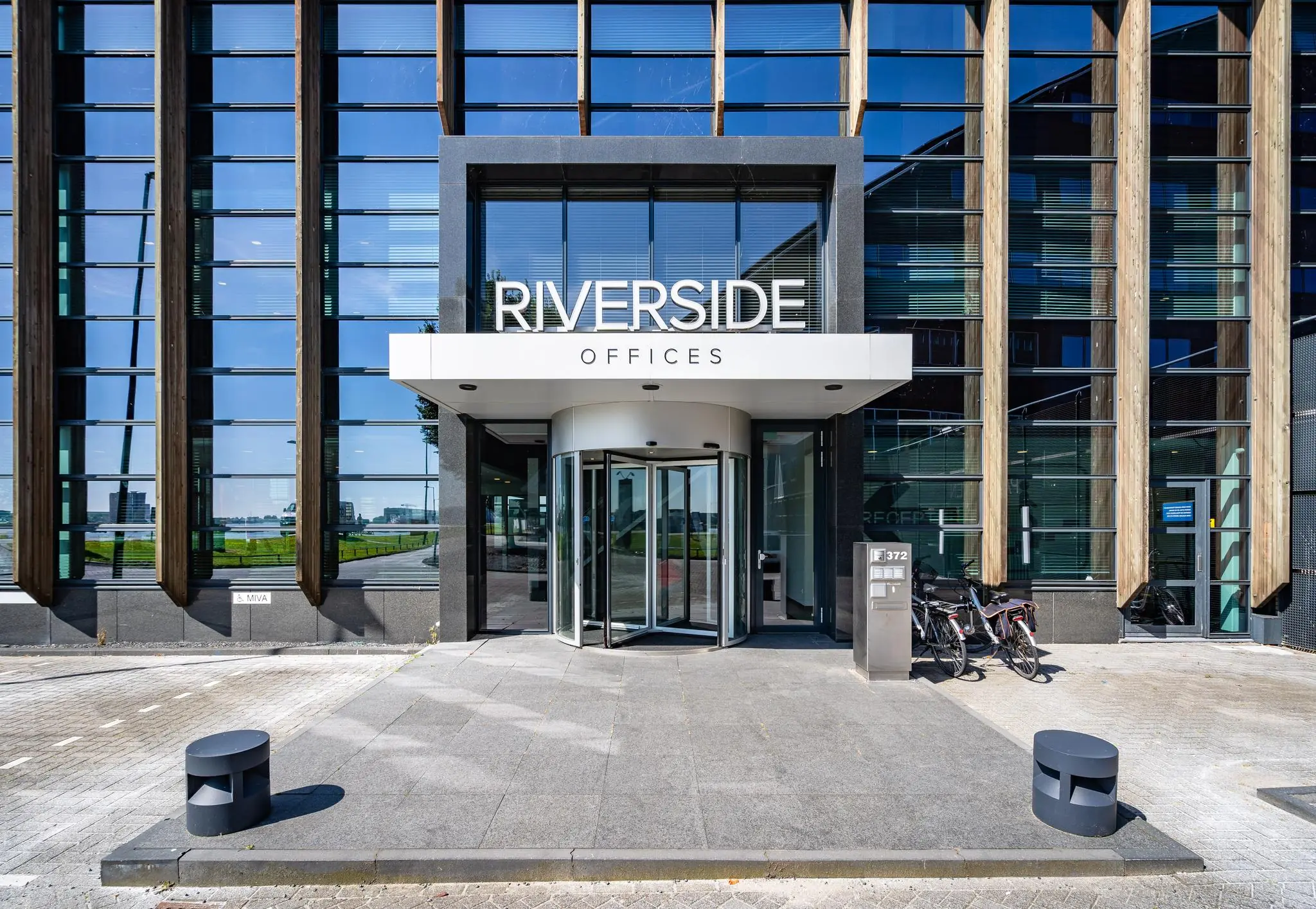 Ingang van het Riverside Offices-gebouw aan de Rivium Promenade met een glazen gevel en draaideur.